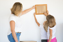 Mother And Daughters Hanging Blank Photo Frame On White Wall. Back View Of Blonde Mom Holding And Aligning Empty Picture With Help Of Two Kids. Family, Relocation And Moving Day Concept
