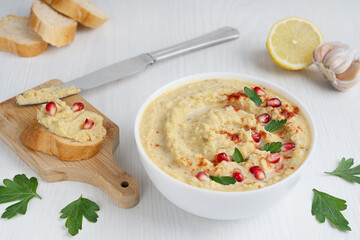 Wall Mural - Vegetarian hummus made of mashed cheakpeas, lemon and garlic decorated with pomegranate seeds and parsley served in bowl with bread slices and knife on white wooden background at kitchen. Horizontal