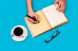 © Mercedes Fittipaldi - Manos de mujer escribiendo en un diario cuaderno junto a una taza de café y unas gafas sobre un fondo azul liso y aislado. Vista superior. Copy space