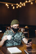 © mykolastock - Stylish smart hipster male wearing protective pandemic face mask sitting in cafe and using smartphone