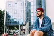 © mykolastock - Cheerful bearded hipster male relaxed sitting in the city, holding smartphone, smiling and looking away