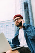 © mykolastock - Serious stylish modern bearded entrepreneur wearing red hat and spectacles working on laptop and talking on phone