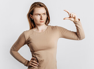 Young beautiful girl frowns and shows small size measure with her hand gesture on a white isolated background. A woman points to an idea, a place for advertising. Negative brunette in a beige jumper.