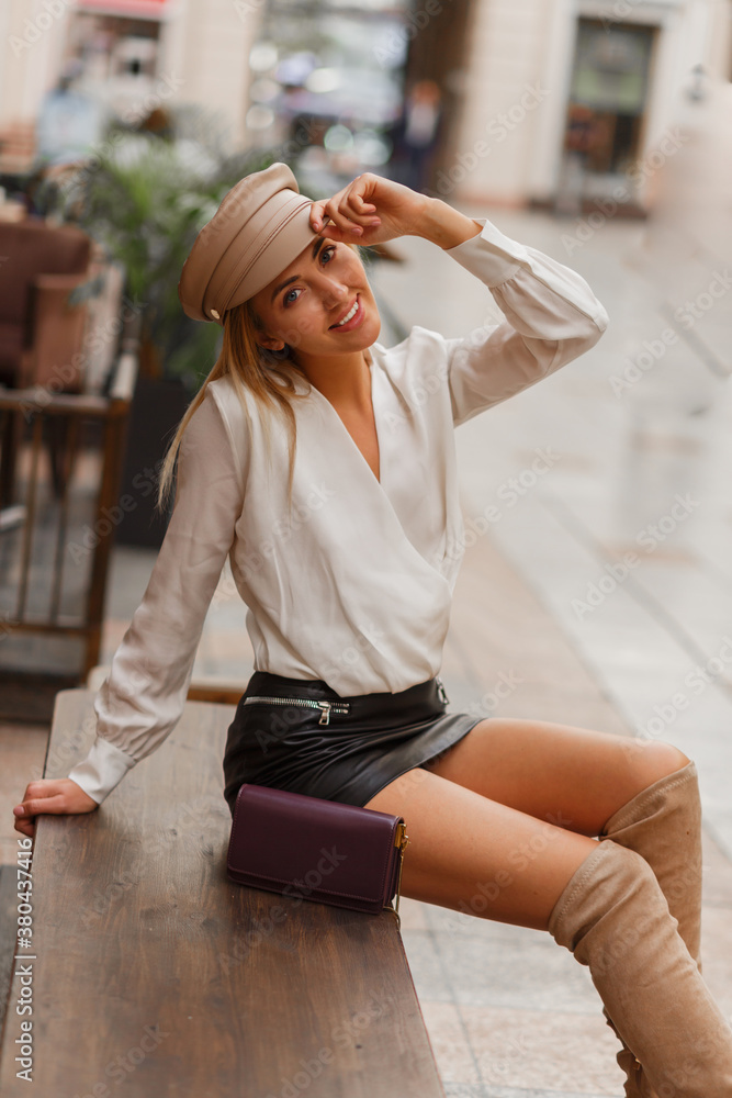 Blond smiling european woman in trendy autunb cap posing outdoor. Wearing white blouse and leather skrt and knitted block  thigh high boots in beige. - obrazy, fototapety, plakaty 