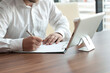 © vadish - Businessman in a white shirt with a digital tablet in his hands signs a contract in the office. Workplace with a cup of coffee and a document with a pen on a wooden table.