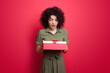 © Strelciuc - Surprised caucasian woman with curly hair wearing dress and holding a gift posing on a red studio wall