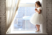 A Beautiful Little Girl In A White Dress Is Waiting For Santa Claus At The Window. Children's Fairy Tales And Dreams. Happy Childhood. Festive Smart Clothes.