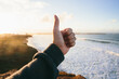 © AlejandroMCB photo & film/Stocksy - Male hand doing a thumbs up in front of beautiful scenic views of a natural sea shore at sunset