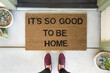 © Amy Covington/Stocksy - Woman standing in front of a doormat reading ___It___s so good to be home__ÔøΩ