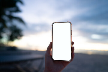 Wall Mural - Woman hand holding mobile phone with blank white screen mockup on the beach in the morning.