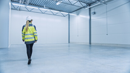Poster - Female Engineer Wearing Safety Hat Walks Across Empty Factory Floor, Inspecting, Planning Machinery Layout. Professional Industrial Designer Strategizing about New Manufacturing Plant Design