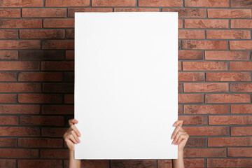 Sticker - Woman holding white blank poster near red brick wall, closeup. Mockup for design