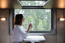 A Young Beautiful Woman In A White Shirt Is Sitting In A Train Compartment With A Cup Of Tea And Looking Out The Window. Travel By Train.