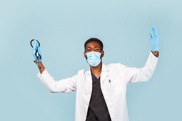 happy African American male doctor with stethoscope wearing mask and gloves on blue background. winner