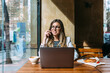 © MyMicrostock/Stocksy - Happy businesswoman using laptop in cafeteria and talking on cellphone