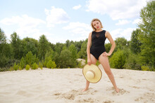 Young Blonde Woman In A Black Swimsuit With A Hat Stands On The Sand