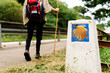 © Alberto Menendez/ADDICTIVE STOCK - Back view of cropped lonely unrecognizable hiker with backpack and trekking stick walking on path near signpost with scallop shell symbol of Camino de Santiago in Spain