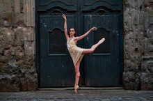 Full Body Side View Of Slim Young Ballerina In Dress And Pointe Shoes Performing Graceful Ballet Pose Against Weathered Door Of Aged Stone Building On Street Of Old City