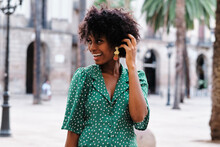 Happy Young African American Female With Curly Hair And Stylish Earrings Wearing Trendy Green Polka Dot Blouse Looking Away While Standing Against Blurred Urban Background