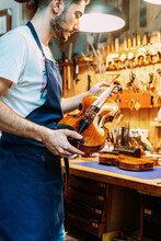 Side View Of Young Craftsman In Apron Standing With Shiny Violin Near Workbench In Modern Workshop