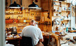 © Javier Ballester/ADDICTIVE STOCK - Back view of unrecognizable male luthier master sitting on chair and catching tool from wall while making instruments in workshop