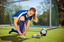 Glad Female Athlete Putting On And Tying Boots Near Ball And Goal While Preparing For Football Training On Field