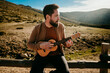 © Lucia Romero/ADDICTIVE STOCK - Serious bearded traveler sitting on fence against cloudy sky and playing ukulele on sunny day in Puerto de la Morcuera, Spain