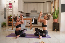 Blonde Mother And Daughter Wearing The Same Sport Clothes Sitting On A Mat On The Kitchen Floor Doing Yoga
