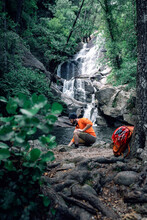 Side View Of Traveler With Backpack Sitting On Rock And Admiring Amazing Landscape Of Green Woods With Fast River