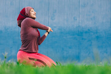Smiling Arab Female Athlete Wearing Hijab Sitting On Green Lawn Stretching Arms With Eyes Closed Before Workout