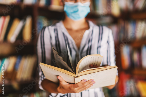 Young college girl with face mask on standing in the library and holding a book during corona pandemic.