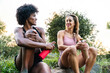 © Luis Manuel Munoz/ADDICTIVE STOCK - Low angle of smiling young multiracial girlfriends in sportswear drinking water and chatting while sitting on stone and resting after workout in nature