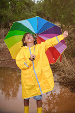 Vertical Photo Of A Blond Boy In A Raincoat With Yellow Water Boots And A Colorful Umbrella In The Rain In A Puddle In The Forest Screaming Happy With His Arm Raised