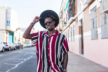 Low Angle Of Confident Young African American Male In Stylish Stripped Shirt And Sunglasses With Hat Looking At Camera While Standing Against Blurred Urban Street