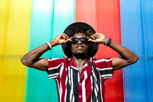 Young African American Male In Striped Shirt And Trendy Sunglasses And Hat Looking At Camera While Standing Against Multicolored Wall