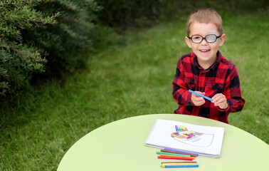 a little boy drawing a picture in the garden (outdoors). wearing glasses and an eye patch (plaster, 