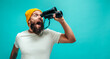 © San4ezz007 - Young bearded hipster guy with binoculars over isolated blue wall wearing white blank t-shirt. Crazy emotions