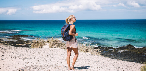 Wall Mural - Caucasian woman with touristic backpack standing at wild shoreline during getaway journey travelling, young hipster girl enjoying summer vacations for visiting Menorca island with turquoise sea