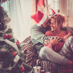 Wall Mural - Concept of real happy life together with caucasian people at home in christmas eve day celebration - cheerful  couple in love with christmas tree - hug and smile for happiness in life together forever