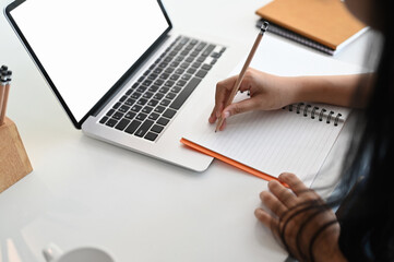 Cropped shot of businesswoman writing her idea on notebook while using laptop with blank screen in office.