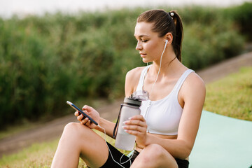 Young slim fit woman with earphones and smartphone listen music in the park at the sunny day