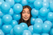 Celebration concept. Stupefied dark haired Asian woman has eyes popped out being on balloon party poses against decorated background. Face of shocked brunette girl around inflated helium balls