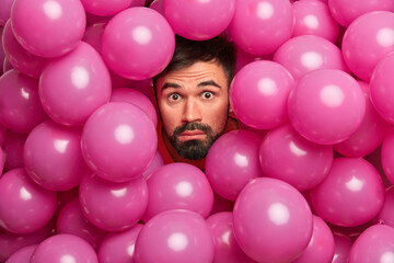 Wall Mural - Impressed bearded European guy poses over decorated photo zone sticks out head through inflated pink balloons has fun on birthday party makes funny shot stares bugged eyes at camera. Celebration time