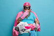 © wayhome.studio  - Indoor shot of stupefied bearded superhero man holds laundry basket covers mouth wears rubber gloves cape and mask stunned to have household duties isolated on blue background. Housework concept