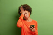 Upset depressed Afro American woman makes face palm as forgot about something important holds mobile phone expresses sorrow and regret blames herself isolated on green background. Negative emotions