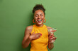 © wayhome.studio  - Overjoyed dark skinned woman laughs happily keeps eyes closed and smiles broadly has fun at party drinks cocktail expresses positive emotions wears yellow shirt poses over vivid green background