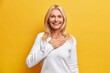 Happy grateful blonde woman in her fifties holds hand on chest and smiles broadly swears be loyal thanks for honesty smiles gladfully dressed in casual white jumper isolated on yellow background