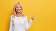 Happy senior woman shows copy space against yellow background smiles pleasantly and has dreamy expression demonstrates vacant advertising spot wears white jumper recommends good offer or discount
