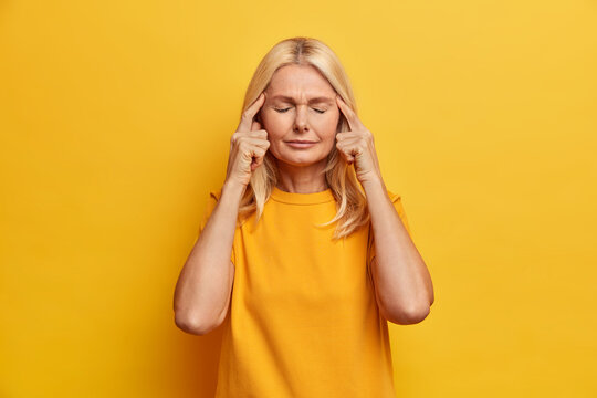 Portrait of attractive senior lady stands with eyes closed keeps fingers on temples thinks about something and tries to find solution has bad memory dressed in casual wear isolated on yellow wall