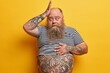 © WHstudio Leushin N - Funny bearded man with fat belly keeps hand on head forgets something very important has thick long beard dressed in undersized striped t shirt isolated on yellow background. Frustrated overweight guy